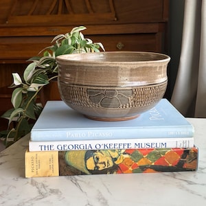 Könnte beinhalten: Eine braune Keramikschale mit einem strukturierten Blumendesign liegt auf einem Stapel von Büchern. Die Bücher tragen Titel wie "Pablo Picasso" und "The Georgia O'Keeffe Museum". Die Schale ist neutralfarben und wirkt handgefertigt.