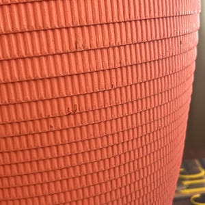 May include: Close-up of a bright orange, corrugated cardboard planter. The planter has a rounded shape and a textured surface.