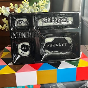 May include: Black ceramic tray with four compartments labelled "Watch", "Pen & Pencil", "Cash", and "Wallet". The tray is labelled "Overnighter".