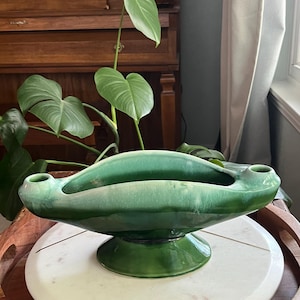 May include: A vintage, green ceramic centerpiece bowl with a pedestal base. The bowl has a unique, elongated shape with two small, cylindrical openings on either side. The glossy finish and varying shades of green add depth to the piece.