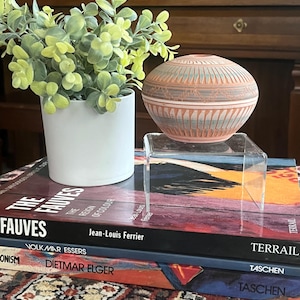 Könnte beinhalten: Ein dekoratives, rundes Töpfergefäß in Terrakotta-Farbe mit geometrischen Mustern steht auf einem transparenten Acrylständer. Daneben befindet sich ein weißer Topf mit einer grünen Pflanze. Die Gegenstände liegen auf einem Stapel Bücher mit farbigen Einbänden.