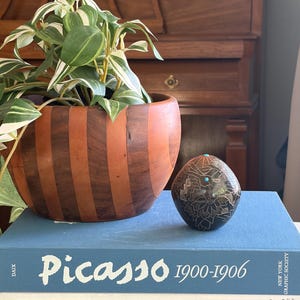 May include: A wooden bowl with alternating dark and light brown stripes holds a green and white plant. A dark, patterned decorative egg with turquoise accents sits on a stack of books. The books are titled "Picasso 1900-1906", "The Georgia O'Keeffe Museum", and "Bonnard".