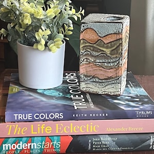 Puede incluir: Un jarrón de cerámica con un diseño ondulado abstracto en tonos marrones, rosas y verdes. El jarrón está colocado sobre tres libros, con los títulos "True Colors", "The Life Eclectic" y "Modernstarts" visibles.
