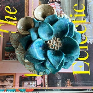 Op de afbeelding: Een keramische bloemenvaas met drie openingen, elk in de vorm van een trompet. De vaas is geschilderd in tinten blauw en groen, met een wit centrum en bruine accenten. De vaas staat op een boek met het woord "Eclectic" op de omslag.