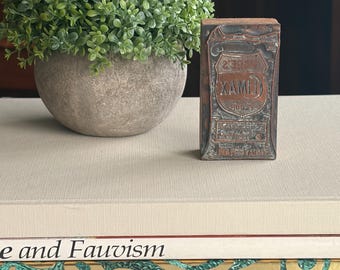 Puede incluir: Un pequeño objeto metálico rectangular y desgastado con el texto "XAMLI" se muestra sobre una superficie beige. Una planta verde y frondosa en una maceta gris está a la izquierda. Libros con los títulos "Fauvismo" y "EVELYN AND JEROME ACKERMAN" están en primer plano.