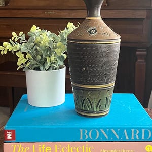 May include: A decorative ceramic vase with a dark brown, textured body and a lighter brown top. The vase has a green base with a geometric design and is sitting on a stack of books. A small potted plant is to the left.