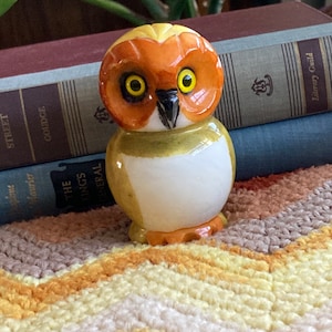 May include: A ceramic owl figurine with orange, yellow, and white paint. The owl has large, round eyes with yellow pupils.