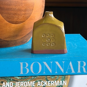 May include: An olive-green ceramic vase with a rectangular body and a narrow neck. The vase has nine circular indentations on the front and a reddish-brown base. A large wooden bowl is in the background. The vase is likely around 10 cm tall.