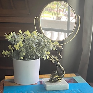 May include: A gold-toned vanity mirror with a round reflective surface. The mirror is supported by a decorative stand featuring a dolphin design, set on a white marble base. A small potted plant with green leaves sits to the left.