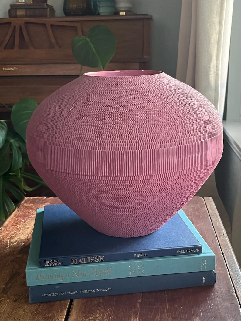 May include: A large, pink, ceramic vase with a textured surface sits on top of a stack of three books. The top book is blue and has the text "MATISSE" printed on the cover. The middle book is teal and has the text "Painting a New World" printed on the cover. The bottom book is blue and has the text "ARCHITECTURAL DIGEST AMERICAN INTERIORS" printed on the cover.