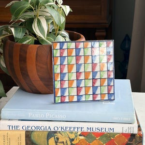 May include: A colorful square tile with a geometric pattern of red, orange, green, and blue triangles. The tile is displayed on a stack of books, including one titled "Pablo Picasso". A wooden bowl with a plant is in the background.