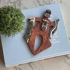 Puede incluir: Un colgante de pared de cerámica marrón con un diseño único, con detalles en crema, verde y dorado. La obra de arte está colocada sobre un libro azul claro, con una pequeña planta verde en el fondo. La pieza tiene una superficie texturizada.