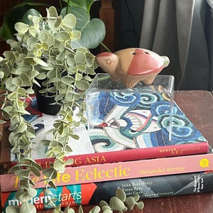 May include: A stack of books with titles "Life in Asia", "The Life Eclectic", and "Modern Starts" with a small terracotta-coloured ceramic animal figurine on top of the books. The figurine is in the shape of a turtle with a red and brown glaze.