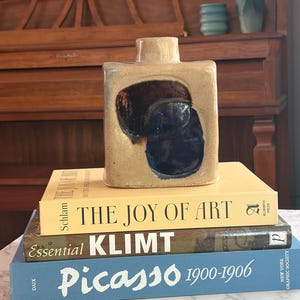 May include: A rectangular ceramic vase with a beige speckled finish and a central dark blue and brown glazed indentation, resting on a stack of three books titled The Joy of Art, Essential Klimt, and Picasso 1900-1906 with visible text.