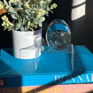 May include: A clear glass paperweight with iridescent blue and green swirls sits on a clear acrylic stand. The stand is on top of a stack of three books. The top book is blue and has the word "BONNAI" printed on the cover. The middle book is pink and has the words "The Life Eclectic" printed on the cover. The bottom book is red and has the words "modernstarts" printed on the cover.