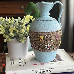 May include: A light blue decorative pitcher with a textured surface and a handle. The pitcher features a decorative band with floral and gold accents. It is sitting on a stack of books, including one titled "MAISON PARISIAN CHIC AT HOME".