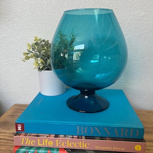 Op de afbeelding: Een turquoise glazen vaas met een brede body en een donkere basis. De vaas staat op een stapel boeken, waaronder een met de titel "Bonnard". Links van de vaas staat een kleine plant in een pot. De boeken staan op een houten oppervlak.