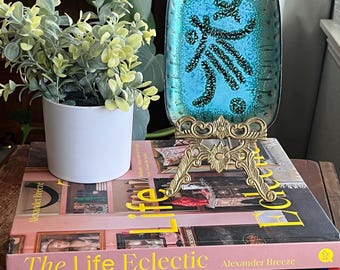 May include: A teal blue glass dish with a black and white abstract design sits on a gold metal stand. The stand is on top of a stack of three books. The top book is pink and has the title "The Life Eclectic" in yellow letters. The middle book is red and has the title "modernstarts" in white letters. The bottom book is a multi-colored book with a brown, gold, blue, and red striped cover.