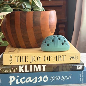 May include: A teal ceramic flower frog with multiple holes sits atop a stack of three books titled The Joy of Art, Essential Klimt, and Picasso 1900-1906. Behind the books is a rounded wooden bowl with vertical stripes and a green leafy plant.
