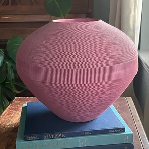May include: A large, pink, ceramic vase with a textured surface sits on top of a stack of three books. The books are blue and teal and have titles that include "Matisse", "Painting a New World", and "Architectural Digest American Interiors".