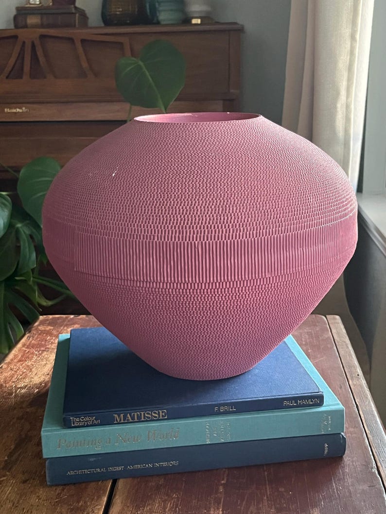 May include: A large, pink, ceramic vase with a textured surface sits on top of a stack of three books. The books are blue and teal and have titles that include "Matisse", "Painting a New World", and "Architectural Digest American Interiors".