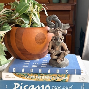 May include: A small, seated clay figurine with a dark grey, braided headdress and circular ear ornaments, positioned on a stack of three books titled Bauhaus, Evelyn and Jerome Ackerman, and Picasso 1900-1906, next to a wooden bowl with a plant.