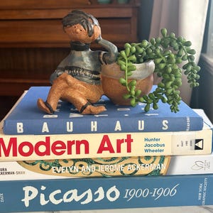 May include: A ceramic figurine of a person sitting next to a small pot containing a green succulent plant, placed on a stack of four books titled Bauhaus, Modern Art, Evelyn and Jerome Ackerman, and Picasso 1900-1906 with blue, white, and red text.