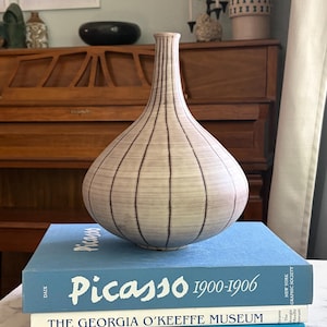 May include: A beige, teardrop-shaped vase with dark brown vertical lines. It rests on a stack of books, including one titled "Picasso 1900-1906". The books are various shades of blue and white.