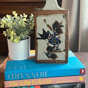 Op de afbeelding: Rechthoekige keramische vaas met een vogel- en bloemenontwerp in zwart, blauw en groen. De vaas heeft een lichtbeige achtergrond en een bruine rand. Hij staat op een stapel boeken, waaronder "BONNARD" en "The Life Eclectic".