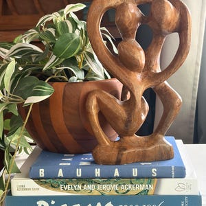 Può includere: Una scultura in legno marrone che raffigura tre figure astratte con arti allungati e superfici lisce, posizionata su una pila di tre libri intitolati Bauhaus, Evelyn and Jerome Ackerman e Picasso 1900-1906, accanto a una pianta.