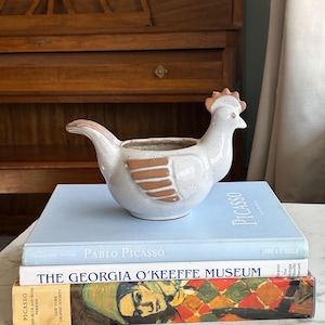 Könnte beinhalten: Ein weißer Keramik-Hühner-Pflanztopf mit braunen Akzenten, der auf einem Stapel Bücher steht. Die Bücher tragen Titel wie "Pablo Picasso" und "The Georgia O'Keeffe Museum". Der Pflanztopf hat eine gesprenkelte Oberfläche und ein detailliertes Design.