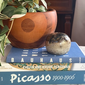 Puede incluir: Un pequeño florero de cerámica esmaltada de forma redonda con acabado moteado en marrón y crema, sobre una pila de tres libros titulados Bauhaus, Evelyn and Jerome Ackerman y Picasso 1900-1906, junto a un cuenco de madera con círculos.