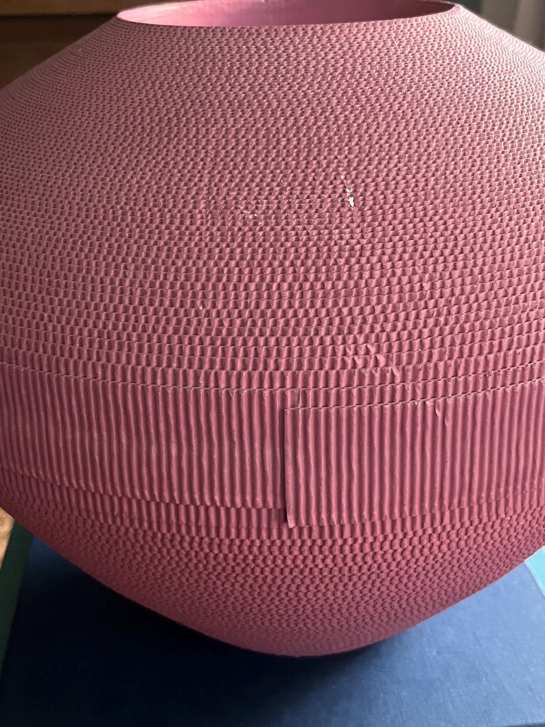 May include: A large, round, pink vase made of cardboard. The vase has a textured surface and is made of multiple layers of cardboard.