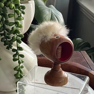 May include: A wooden nutcracker with a white fur hat and a red and brown face. The nutcracker is standing on a clear acrylic stand.