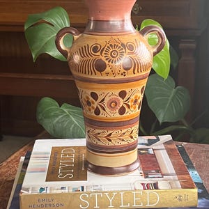 May include: A brown ceramic vase with a floral design in shades of orange, yellow, and brown. The vase has two handles and a rounded base.