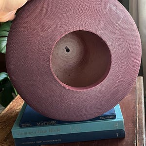 May include: A large, round, purple-colored hat made of woven material. The hat is sitting on top of a stack of three books. The top book has the title "Matisse" and the author "Paul  Hamlvn". The middle book has the title "Painting a New World". The bottom book has the title "Architectural  Art  African  Interiors".