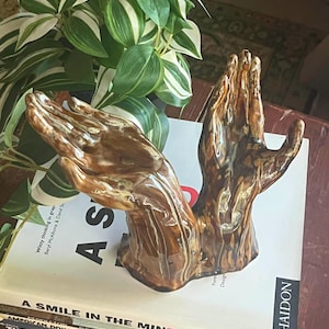 Op de afbeelding: Een bruin en tan keramisch beeldhouwwerk van twee handen in een gebedspositie. Het beeldhouwwerk staat op een stapel boeken, met een groen-witte plant op de achtergrond. Het beeldhouwwerk heeft een glanzende afwerking.