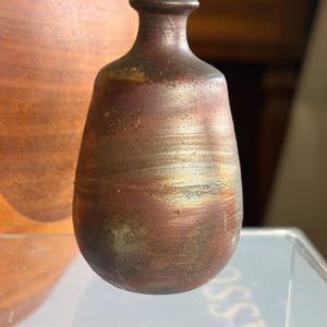 Puede incluir: Un pequeño jarrón de cerámica hecho a mano con cuerpo redondeado y cuello estrecho. El jarrón tiene una rica paleta de colores terrosos, con tonos marrones, dorados y toques de verde. La superficie tiene un aspecto texturizado y orgánico, sugiriendo una creación artesanal única.