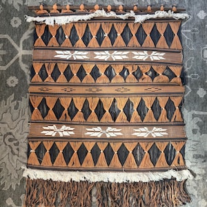 May include: A rectangular wall hanging featuring a dark brown and black woven pattern with rows of knotted orange yarn triangles. The piece includes white embroidered star-like motifs, a wooden top rod, and white and brown fringe at the ends.