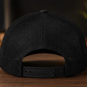 May include: A solid black baseball cap featuring a textured mesh back panel and a matching black plastic snapback closure at the rear. The cap has a structured crown with a central button at the top and a consistent dark colour throughout the design.