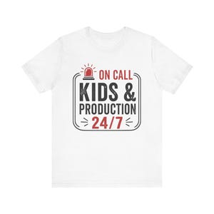 May include: White t-shirt featuring a black and red graphic. The graphic says "ON CALL KIDS & PRODUCTION 24/7" with a red siren icon. The shirt is made of a soft material.