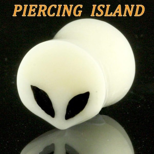 May include: White acrylic ear gauge with a black alien face design. The gauge is a rounded shape with a flat front.