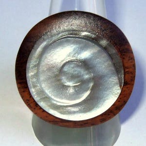 May include: A round wooden ring with a white mother-of-pearl inlay in the center. The inlay has a spiral design.