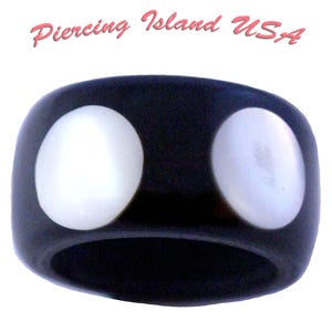 May include: A black ring with two large, oval, white inlays. The ring is smooth and glossy, with the text "Piercing Island USA" in pink script above it. The ring is a bold, eye-catching piece of jewelry.