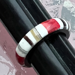 May include: A colorful ring with a striped design featuring red, white, brown, and gray bands. The ring is set against a black background, highlighting its vibrant colors and unique pattern. The ring is a piece of jewelry.