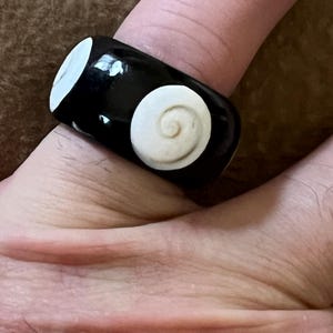 May include: A black ring with two white spiral designs. The ring is worn on a finger and has a glossy finish. The spiral designs are circular and raised, adding texture to the ring.