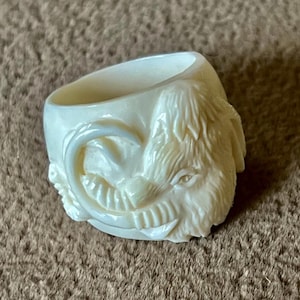 May include: Ivory-colored ring with a carved animal face design. The ring features a detailed depiction of a creature with fur, eyes, and teeth. The ring is set against a brown, textured background.
