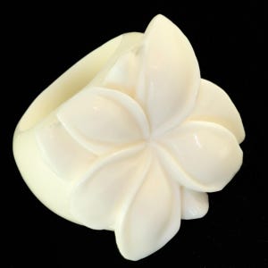 May include: Ivory-colored bangle bracelet featuring a sculpted plumeria flower design. The flower has multiple petals and is the focal point of the bracelet. The bracelet is made of a smooth, glossy material and is set against a black background.