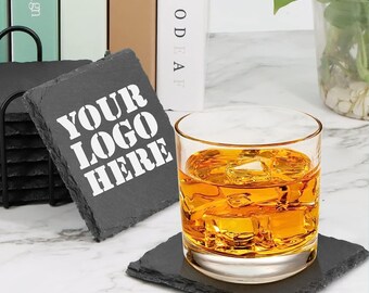以下が含まれることがあります： 黒いスレートコースターのセットで、白い文字で「YOUR LOGO HERE」と書いてあります。アンバー色の液体の入ったグラスが、コースターの1つの上に置かれています。