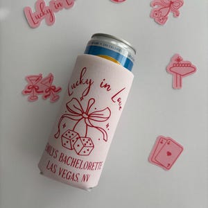 Pode incluir: Um refrigerador de latas branco com fundo rosa e texto vermelho que diz "Lucky in Love" com um la&ccedil;o e dados. O texto abaixo diz "Emily's Bachelorette Las Vegas, NV".